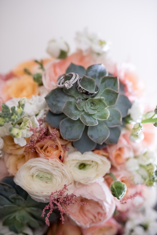 Close up shot of rings on bride's pink, peach, cream, and green bouquet with succulents