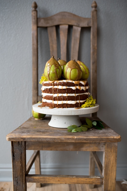 Naked cake topped with caramel and pears sits on a cement pedestal on top of a rustic chair.