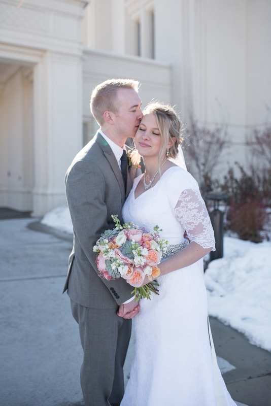 Groom in grey suit kissing bride in white lace gown on temple of forehead