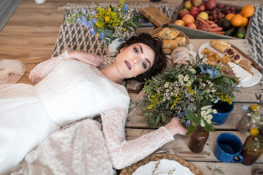 Bride in white lace gown with long sleeves lays on picnic table filled with wildflowers and rustic items.