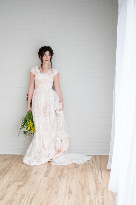 Bride in blush colored dress with lace overlay holds wildflowers and stares into distance