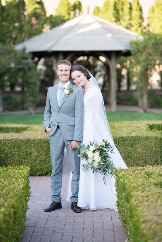 Groom in gray suit and bride in lace gown and long veil with bouquet, standing in the garden at Thanksgiving Point. Bridal session by Flying Gull Photography.